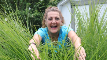 Camille L. Miller smiling joyfully, reaching through tall grass in a bright blue shirt, surrounded by nature.